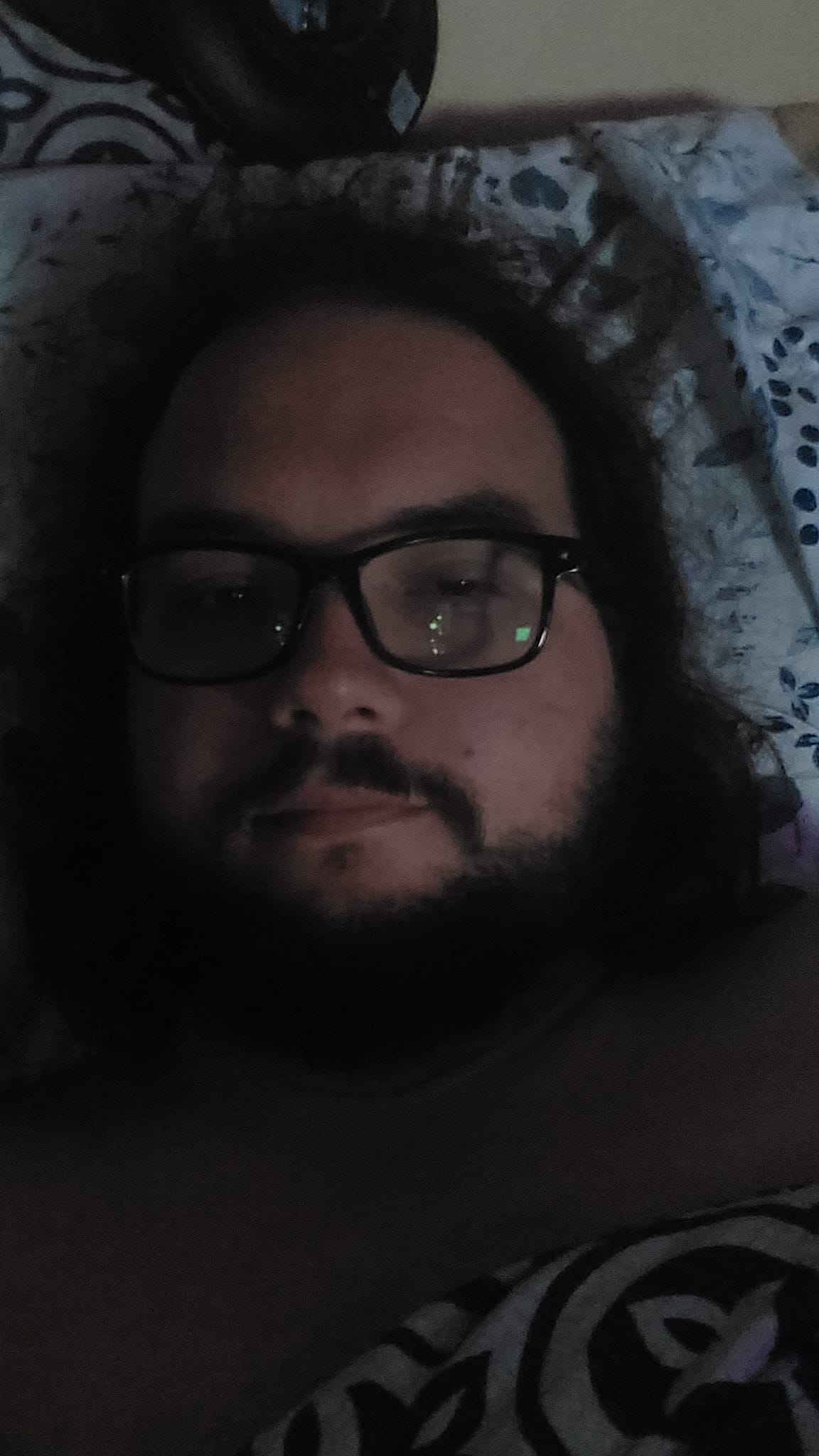 A man with long dark hair and a beard is in bed, the lighting is bad, photo 1